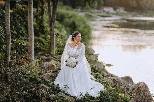 A beautiful bride in a white dress with a bouquet of roses sits on the stones in the garden at sunset against the background of the river, lake. Wedding photography, portrait.