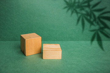 two brown precious wood boxes, blocks, a cubic podium, a branch with canbis leaves on a green...