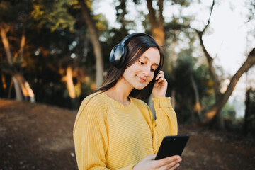 Teenage girl wearing yellow sweater, using headphones for playing music on her smartphone in the park