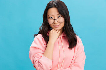 Fototapeta premium Cutie Asian student young lady in pink hoodie sweatshirt wear round glasses smiles at camera posing isolated on blue studio background. The best offer for ad. Eyewear for vision correction concept