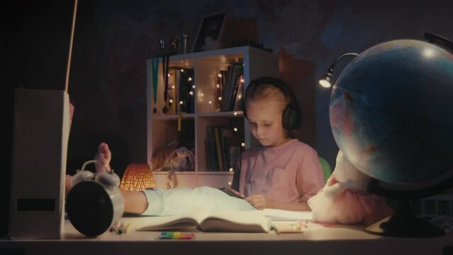 Little cute girl sits with her feet up on the table, playing video games on digital tablet and listening music in headphones at night. Child on social media