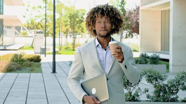 Positive handsome brazilian or hispanic curly haired man, ceo, top manager, real estate agent, holding laptop and glass of coffee, walking outside near business center, drinks coffee, looks away,smile