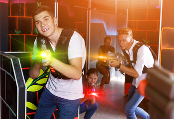 Fototapeta na wymiar Young smiling guys and girls in vests and with laser pistols playing laser tag game in room