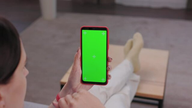 Young woman using smartphone with green mock-up screen indoors of cozy home. Scrolling through social media or online shop, swiping photos or pictures, surfing Internet,watching content videos blogs.