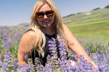 Pretty blonde woman sits in a field of lupine wildflowers, posing with sunglasses and smiling