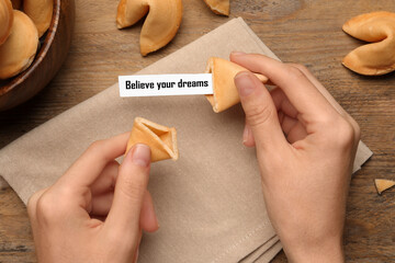 Woman holding tasty fortune cookie with prediction Believe your dreams at wooden table, top view