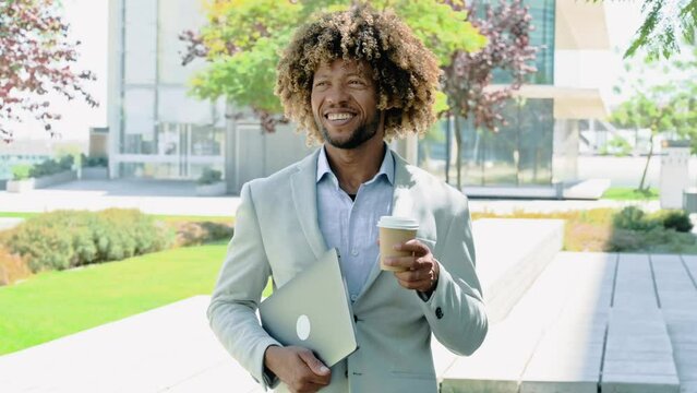 Video in motion of successful brazilian or latino curly man, top manager, real estate agent, holding laptop and glass of coffee, walking outside near business center, drinks coffee, looks away, smile