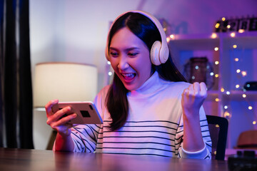 Excited Young Asian woman wearing headset and playing online game on smartphone with live...