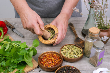 Woman's hands grind cardamom in a wooden mortar. Kitchen gray table with various spices.