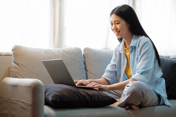 Young asian woman smiling and using laptop while sitting on couch at home.