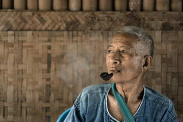 Portrait of an old man wearing traditional clothes and happily smoking tobacco leaves with an ancient wooden pipe.