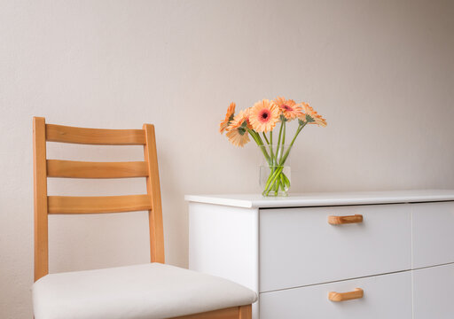 Angled view of orange gerbera daisies in glass vase on white cabinet with chair against beige wall (selective focus)