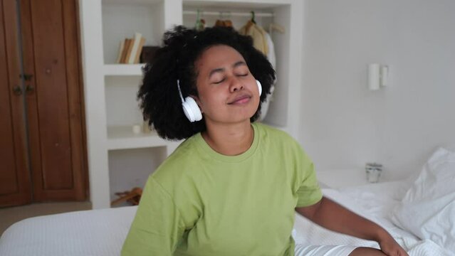 Afro Asian woman listening to music with headphones in the bedroom during quarantine. A mixed-race woman with an Afra hairstyle moves to the beat of the music from her headphones.
