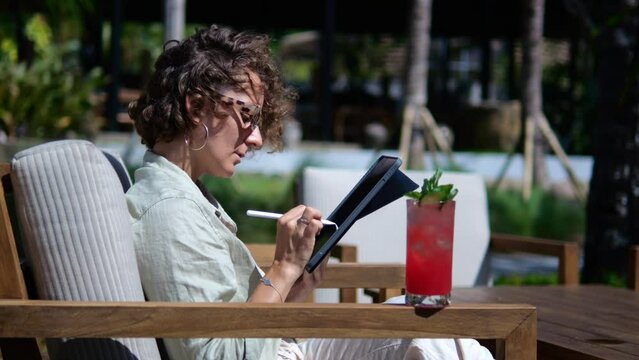 A beautiful girl in sunglasses working as a freelancer sits in a street cafe with a cold drink is engaged in working remotely via the Internet using a graphics tablet to work as an illustrator.