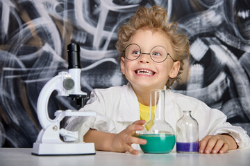 An enthusiastic cheerful boy in lab coat poses at a table with flasks and microscope against the background of blackboard, looking into the camera. Joyful child with hair on end is engaged in science