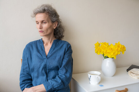 Middle aged woman with grey hair seated next to cup and flowers, looking away from camera with sad expression (selective focus)