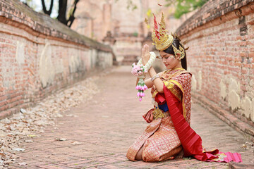 Young fashion and beautiful asian woman in red Thai traditional costume pay respect sawasdee symbol while holding jasmine garland standing in ancient buddha temple Ayutthaya, Thailand. Travel concept