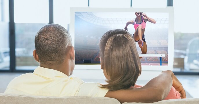Composition of rear view of sports fan couple sitting on couch watching athletics event on tv