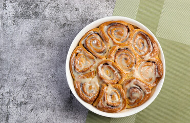 Cinnamon rolls or cinnabon, homemade sweet traditional dessert buns with white cream sauce in a white baking dish on a dark grey background. Top view, flat lay