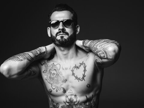 Portrait of handsome confident stylish hipster lambersexual model. Sexy modern man. Naked torso with tattoos.Fashion male posing in studio on dark background in sunglasses. Black and white