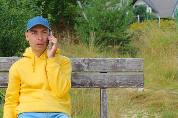 A young man in a cap is talking on the phone while sitting on a wooden bench in nature in the...