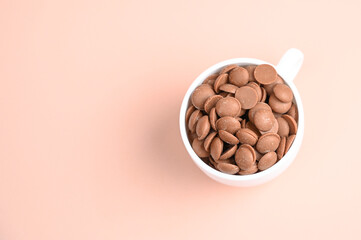 Chocolate callets in a white cup on a pastel background with copy space. Tempered chocolate confectionery concept