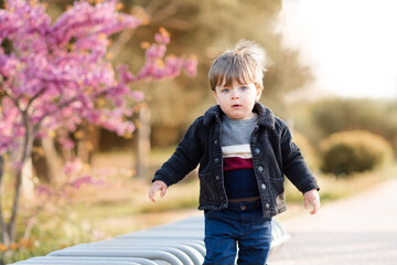 Cute baby boy 1-2 year old wear casual denim jacket and pants posing in park outdoor over nature background. Stylish little child walk on city street. Childhood. Autumn season.