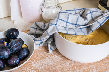Plum on plate, yeast dough in bowl with towel, glass with sugar and paper kitchen towel on wooden kitchen board .