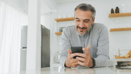 Attractive Middle-aged man using mobile phone or smartphone while standing in kitchen at home.