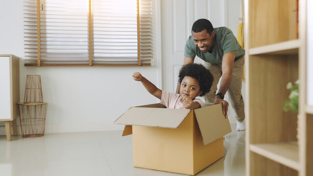 Happy playful African American father and son having fun sitting in cardboard on moving day. Black African man playing with his little boy riding in box.