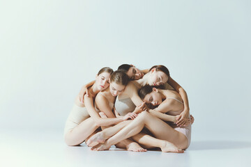 Group of young girls, ballet dancers sitting and hugging, posing isolated over grey studio...
