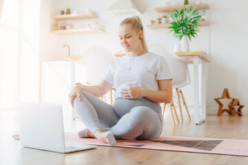 Young pregnant blonde woman practicing online yoga at home with laptop background white kitchen