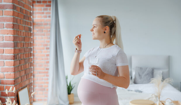 Happy young pregnant woman with white round pill and glass of water in hand, daily vitamins for hair, skin and health of baby