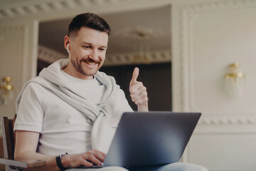 Happy young man freelancer in earphones showing thumb up while talking by video chat with colleague at home office