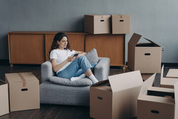 Female orders moving service using phone app on relocation day sitting in room with carton boxes