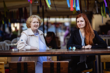 Two women together - blonde in light and redhead in dark clothes talking at the bar of a street cafe. Photo shoot based on the series 