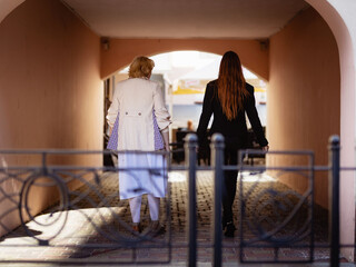 Two women together - blonde in light and redhead in dark clothes walk together on the background of the arch, back view. Cosplay - female version of Angel and Demon (Good Omens)