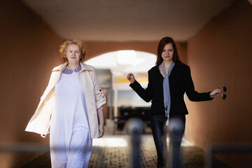 Two women together - blonde in light and redhead in dark clothes walk on the background of the arch. Cosplay - female version of Angel and Demon (Good Omens)