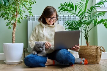 Middle-aged woman in warm clothes with cat using laptop at home
