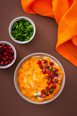 Pumpkin puree soup of butternut squash in bowl with pomegranate seeds and green onions, top view on plate with food on brown background