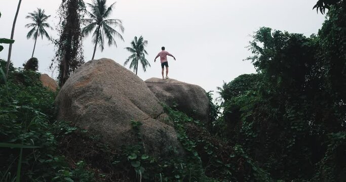 View from the back of young athletic man tourist in pink shirt and casual shorts running up on a rock, contemplates an exotic rainforest nature. He raises his arms up and breathes fresh mountain air