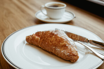 A croissant on a white plate and a cup of black coffee, cutlery on a wooden window-will