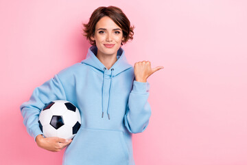 Photo of young adorable gorgeous nice pretty girl wear blue hoodie hold football ball finger pointing empty space positive recommend isolated on pink color background