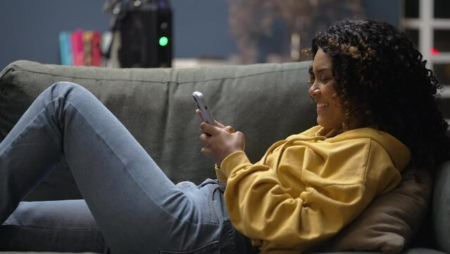 Happy African American girl checking cellphone laid on couch at home living room. One black young hispanic latina woman using phone