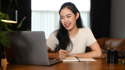 Smiling young woman in casual wear working online, checking email on laptop at home