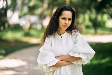 Angry emotions of a curly-haired brunette girl with crossed arms in the park. High quality photo