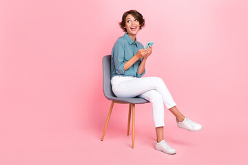 Photo of funny lady trendy outfit sit chair look interested empty space hand hold use telephone gadget isolated on pink color background