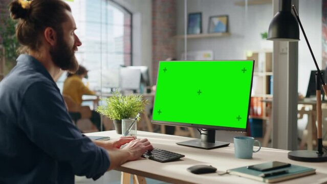 Stylish Long-Haired Bearded Specialist Sitting at a Desk in Creative Agency. Young Stylish Man Working on Desktop Computer with Green Screen Mock Up Display. Office Team Members at Work in Background.