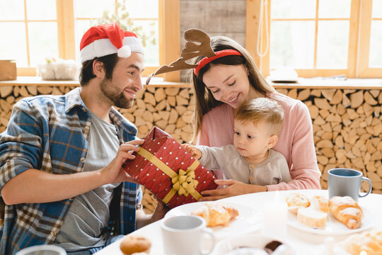 Merry Christmas and Happy New Year! Young caucasian family in Santa`s hats parents giving present gift to little toddler child kid son daughter at home at the table