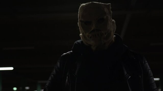 Masked maniac hunts victim at night on doomsday night. Evil scary scarecrow with bag on his head in dark outfit walks through night parking lot and swings baseball bat. Halloween horror.